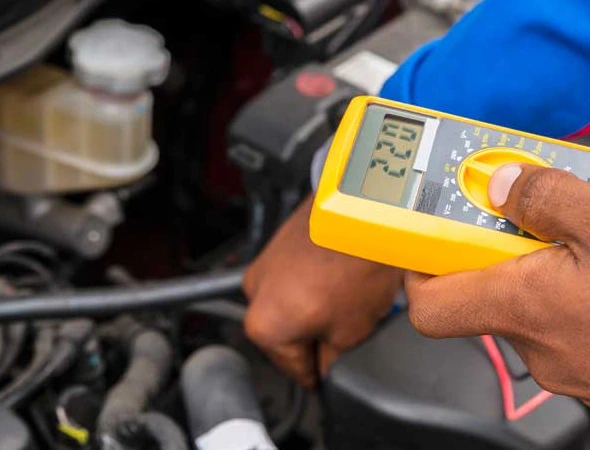 A person conducts inspection and preventative maintenance on a car, holding a multimeter to check electrical systems.